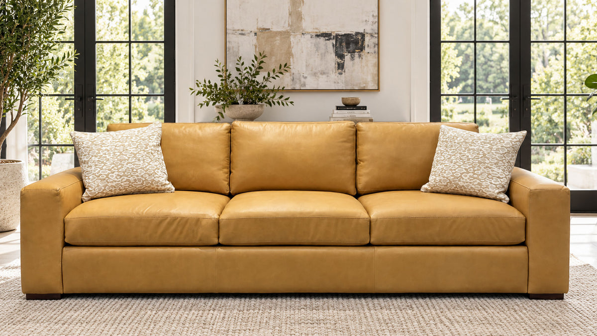 A mustard yellow leather three-seater sofa with two textured cream throw pillows, set in a bright living room with large black-framed windows and a neutral rug.