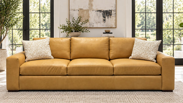 Un sofá de tres plazas de piel de color amarillo mostaza con dos cojines decorativos de color crema con textura, situado en un salón luminoso con grandes ventanas con marcos negros y una alfombra de tonos neutros.