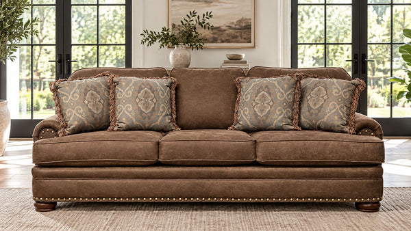 A brown upholstered loveseat with four patterned throw pillows, shown in a bright living room setting with large windows and a rug.