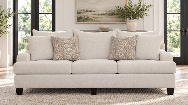 Un sofá de dos plazas tapizado en beige claro, con cuatro cojines de respaldo a juego y dos cojines decorativos con un estampado geométrico en tonos beige y gris, situado en un salón luminoso con grandes ventanas con marcos negros.