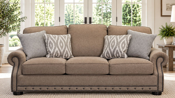 Un sofá de dos plazas tapizado de tres asientos, en un cálido tejido de color beige topo, decorado con cuatro cojines decorativos (tres lisos de color topo y uno con estampado geométrico en gris y blanco). El sofá presenta un ribete de clavos de madera oscura a lo largo de la base y en los reposabrazos redondeados, y se encuentra en un salón luminoso y moderno con grandes ventanas y plantas.
