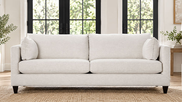 Un sofá de dos plazas tapizado en beige claro, con dos cojines de respaldo, dos cojines de asiento y dos cojines decorativos a juego, que se muestra en un salón luminoso con ventanas de marcos negros y una alfombra tejida.