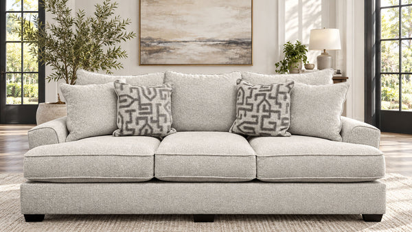 Un sofá de tres plazas tapizado en gris claro en un salón luminoso, decorado con tres cojines decorativos estampados. La estancia cuenta con una alfombra de tonos neutros, una mesita auxiliar con una lámpara, una planta en maceta y un gran cuadro abstracto colgado en la pared.