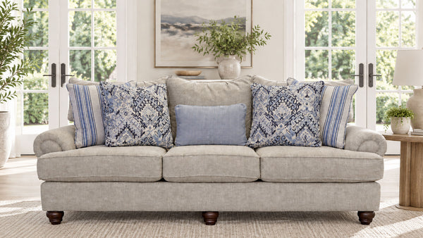 Un sofá de dos plazas tapizado en beige claro con patas de madera oscura, decorado con cuatro cojines decorativos con motivos azules y blancos, situado en un salón luminoso y moderno con grandes ventanas y plantas.