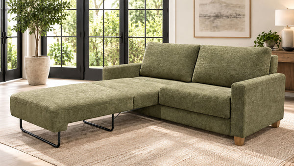 Un sofá modular de color verde oliva con chaise longue sobre una alfombra tejida de color beige, en un salón luminoso y moderno con grandes ventanas con marcos negros.