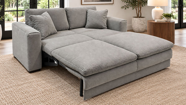 Un sofá chaise longue de tela gris claro con cojines a juego, fotografiado en un salón real con una alfombra tejida, una mesita auxiliar, una lámpara y ventanas al fondo.