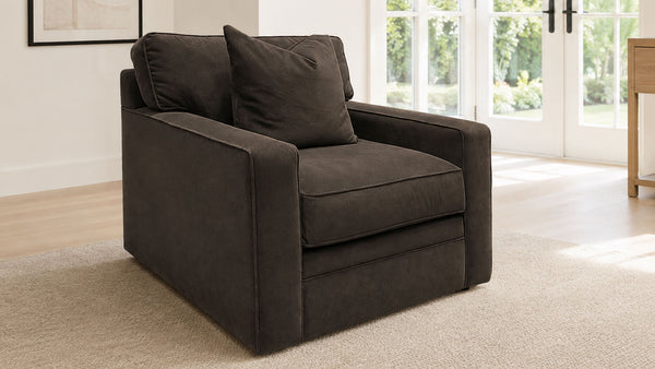Un sillón tapizado de color marrón oscuro con un cojín decorativo a juego, situado sobre una alfombra beige en un salón luminoso y moderno con grandes ventanales.