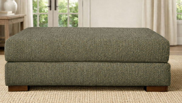 Otomana tapizada en verde en una habitación con ventana y cortinas.