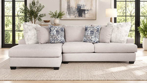 Un sofá modular de color beige claro con una chaise longue orientada hacia la izquierda, decorado con varios cojines decorativos en un salón luminoso y moderno.