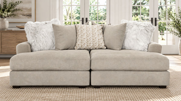 A light beige upholstered sectional sofa with two chaise ends, styled with five throw pillows including two white faux fur pillows and two textured beige pillows, set in a bright living room.