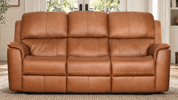 A three-seat power reclining loveseat sofa upholstered in warm tan leather, shown in a staged living room setting with a woven rug and window background.