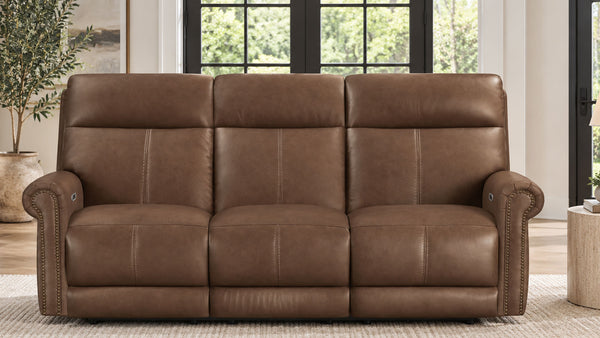 A three-seat power reclining loveseat sofa upholstered in brown leather, shown in a modern living room setting with large windows and a light-colored rug.