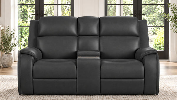 A black leather reclining loveseat sofa with a center console armrest, shown in a bright living room setting with large windows and plants.