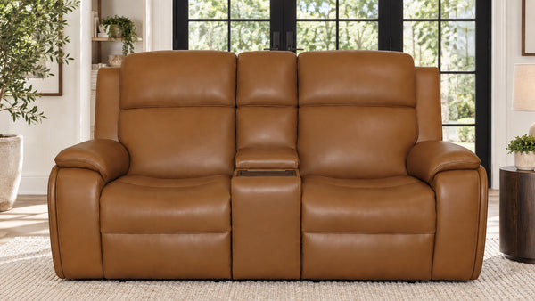 A three-seat power reclining loveseat in smooth tan leather, positioned in a bright living room setting with large black-framed windows and plants in the background.