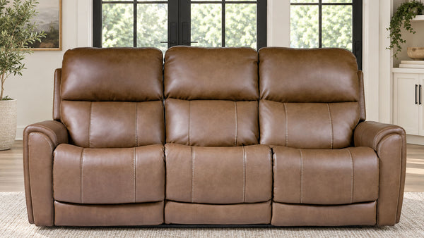 A three-seat power reclining loveseat sofa upholstered in brown faux leather, positioned in a bright living room setting with windows and plants in the background.
