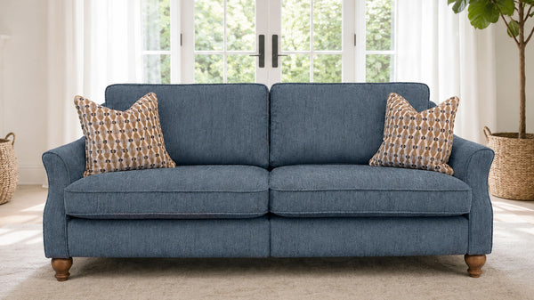 A two-seater loveseat sofa upholstered in textured blue fabric, styled with two patterned throw pillows and wooden legs, set in a bright living room with a window and potted plant in the background.