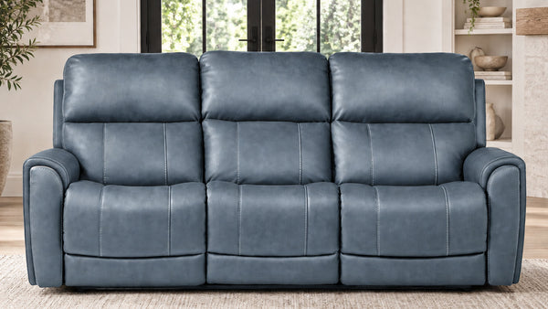 A three-seat reclining loveseat sofa upholstered in textured blue faux leather, positioned in a modern living room setting with a light beige rug, shelving, and windows in the background.
