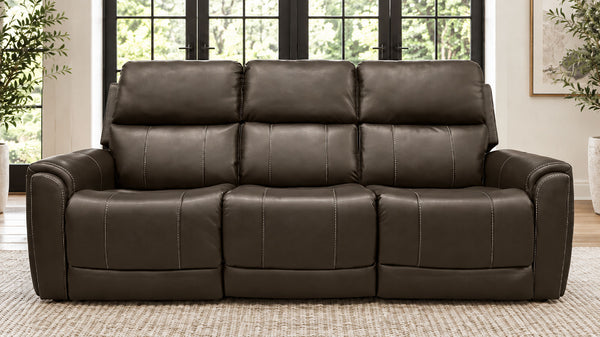 A three-seat reclining sofa upholstered in dark brown leather, positioned in a bright living room setting with a textured rug and large windows in the background.