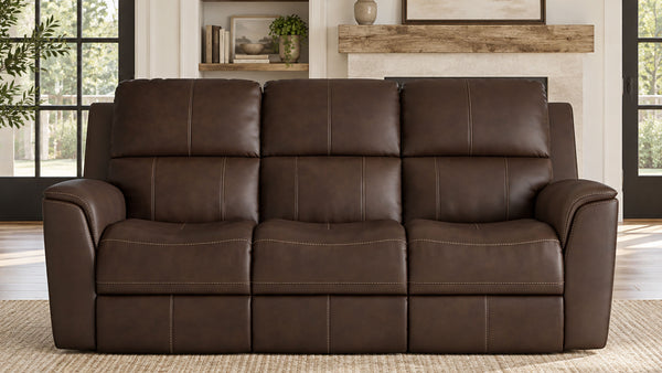 A three-seat power reclining loveseat sofa upholstered in dark brown leather, positioned in a modern living room setting with a fireplace and large windows in the background.