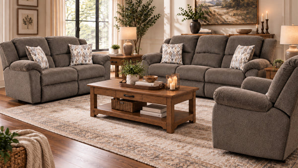 Un salón decorado con un sillón reclinable tapizado en gris, un sofá de dos plazas y un sillón, dispuestos alrededor de una mesa de centro de madera sobre una gran alfombra estampada. La estancia cuenta con ventanas, lámparas, mesitas auxiliares, plantas y cuadros.