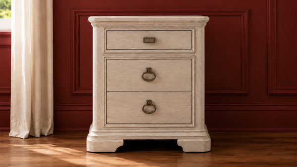 Una mesita de noche de tres cajones con un acabado color crema envejecido y tiradores circulares de metal oscuro, que destaca sobre una pared panelada de color rojo oscuro y un suelo de madera.