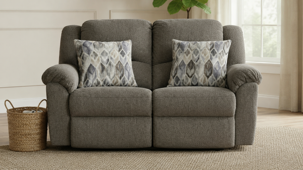 A gray upholstered reclining loveseat with two patterned throw pillows, shown in a bright living room setting with a woven basket on the floor and a window in the background.