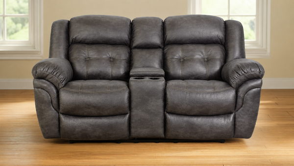 A dark gray faux leather reclining loveseat with a center console cup holder, set against a background of beige walls, windows, and a light wood floor.
