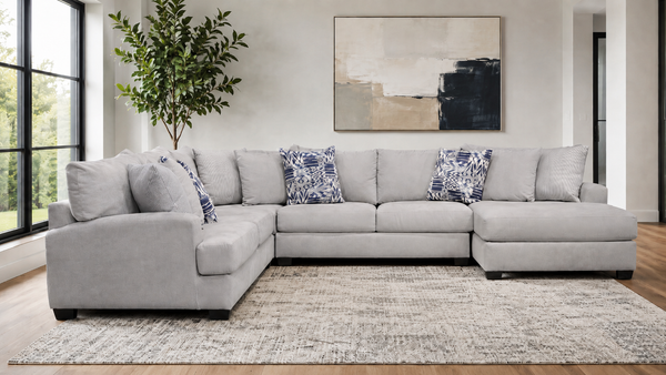 A light gray sectional sofa with matching throw pillows and two blue-and-white patterned accent pillows, staged in a bright living room with a rug, wall art, and a plant.