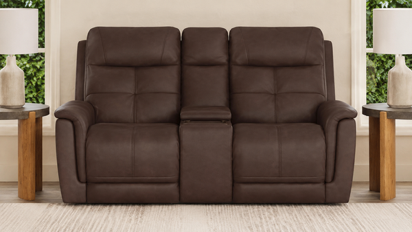 A three-seat power reclining loveseat in dark brown faux leather, positioned in a living room setting with side tables, lamps, and windows in the background.
