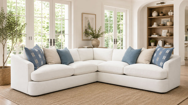 Un sofá modular blanco con cojines decorativos azules y beige, situado en un salón luminoso y moderno con grandes ventanales, una alfombra tejida y una estantería de madera al fondo.