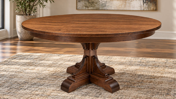 A round wooden dining table with a dark brown pedestal base, shown in a staged dining room setting on a textured rug with hardwood floors and a potted plant in the background.