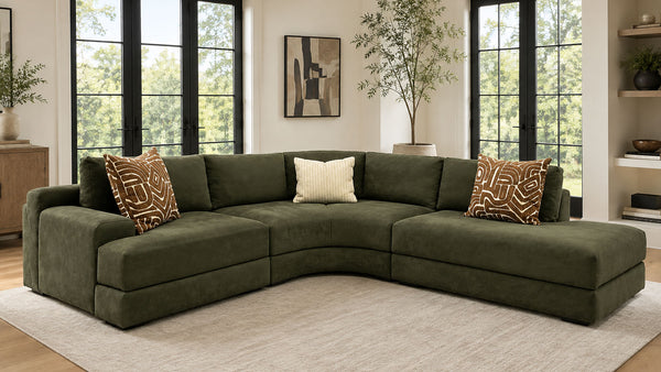 Un sofá modular de color verde oliva con dos cojines decorativos de motivos en tonos marrones y crema, situado en un salón moderno con grandes ventanas con marcos negros y una alfombra de color beige claro.