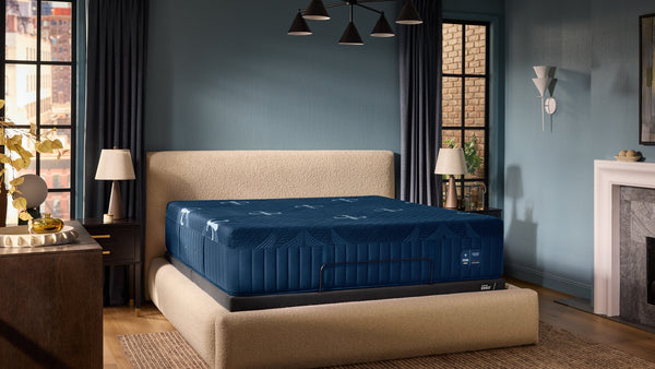A luxury mattress on a matching upholstered platform bed frame, styled in a modern bedroom with blue walls, dark curtains, and warm wood flooring.