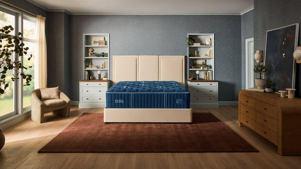 A modern bedroom featuring a bed with a cream upholstered headboard and footboard, a blue striped mattress, and a dark brown rug on a light wood floor. The room has textured blue-gray walls, built-in white shelving, a wooden dresser, and a window with sheer curtains.