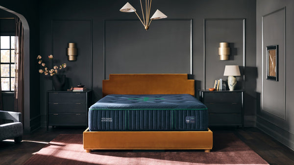 A luxury mattress shown in a styled bedroom setting. The mattress has a dark blue top and a ribbed navy blue side with a green logo, set on a mustard yellow platform bed frame.