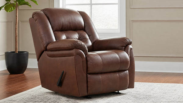 A brown micro recliner chair with a tufted backrest and padded armrests, shown in a staged living room setting with a window, wood floor, rug, and potted plant.