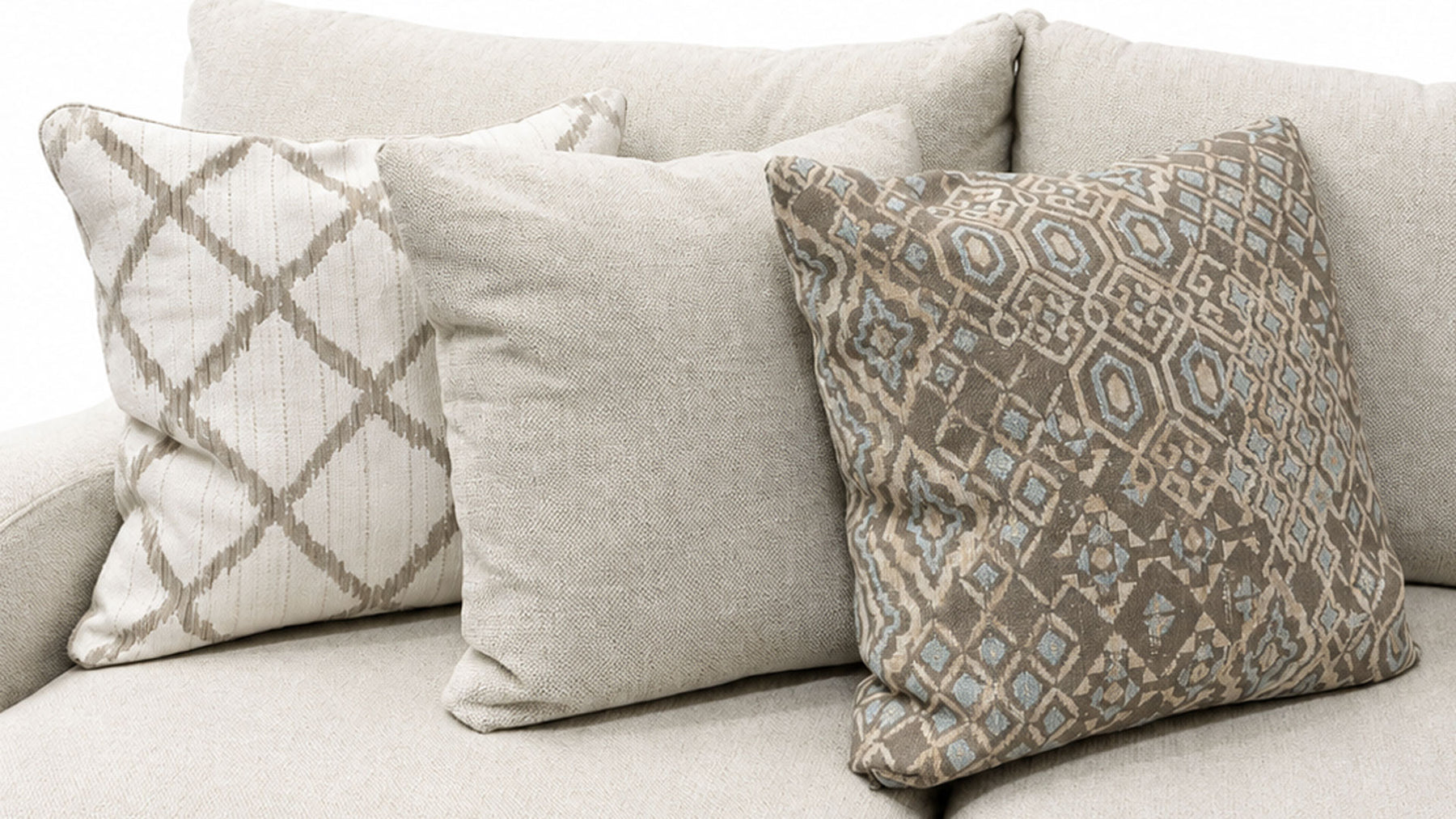 Three decorative throw pillows arranged on a light beige sofa. The center pillow is a solid light beige linen texture, flanked by two patterned pillows in beige, taupe, and light blue-gray tones.