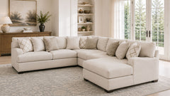 A light beige sectional sofa with a chaise lounge in a bright, modern living room. The sofa is styled with multiple neutral-toned throw pillows, set on a large gray patterned area rug.