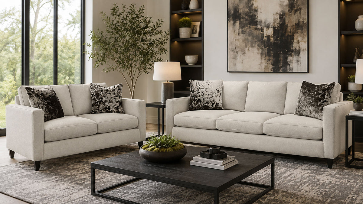 A staged living room featuring a light beige upholstered loveseat and matching sofa, styled with black and white patterned accent pillows, a dark wood coffee table, and a large abstract wall art piece.