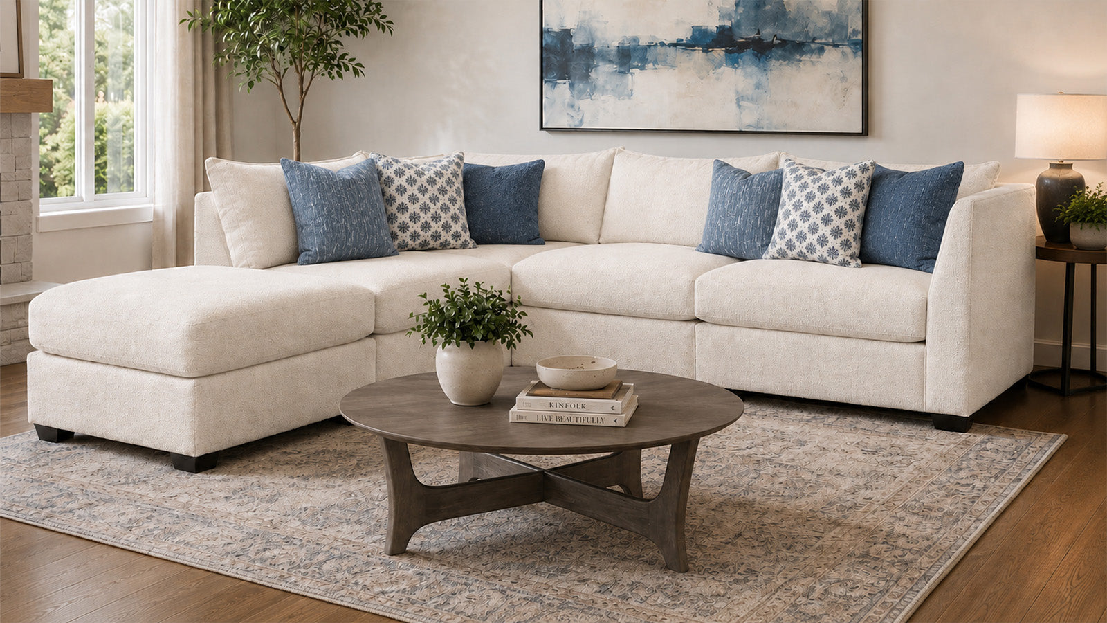 Un sofá modular de color crema con cojines decorativos azules y estampados, dispuesto en un salón luminoso con una mesa de centro redonda de madera, una alfombra estampada y un gran cuadro abstracto en la pared.