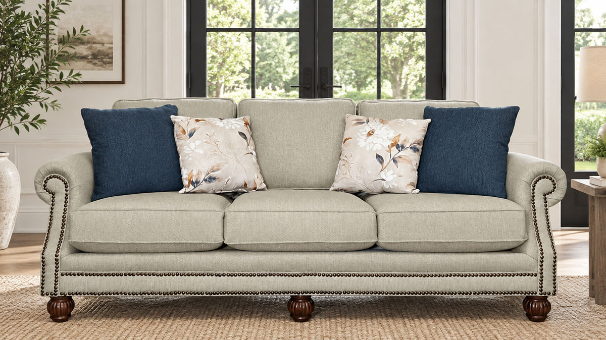 A three-seat upholstered sofa in a light beige fabric with rolled arms, nailhead trim, and dark wood legs, styled with four throw pillows in a bright living room.