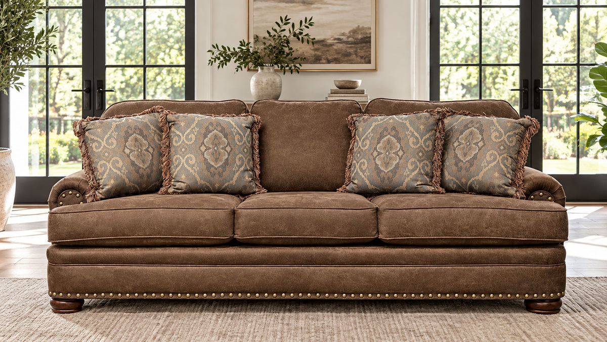 A brown upholstered loveseat with four patterned throw pillows, shown in a bright living room setting with large windows and a rug.