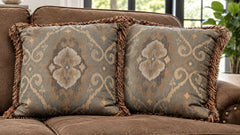 Two decorative throw pillows with a blue-gray and beige damask pattern and brown fringe, resting on a brown sofa in a living room setting.