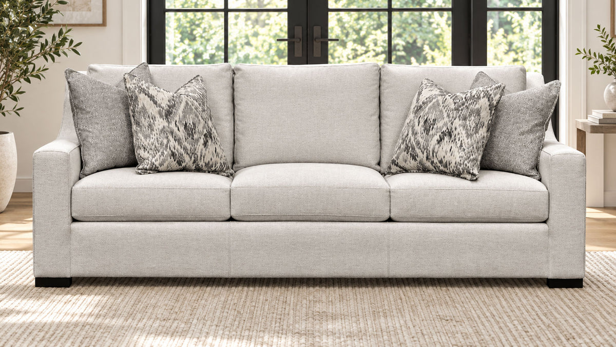 Un sofá de tres plazas tapizado en beige claro con cuatro cojines decorativos, entre los que se incluyen dos cojines con estampado gris y dos cojines de color gris liso, que se muestra en un salón luminoso.