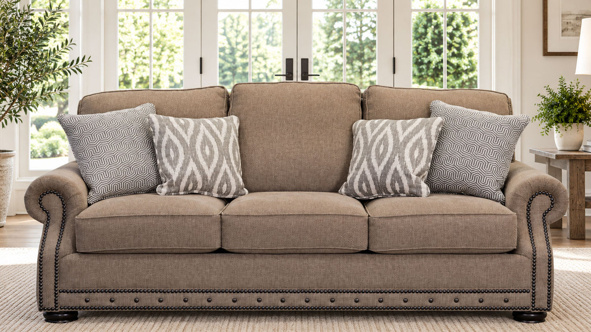 A three-seat upholstered loveseat in a warm taupe-beige fabric, styled with four throw pillows (three solid taupe and one gray-and-white geometric patterned pillow). The loveseat features dark wood nailhead trim along the base and rolled arms, set in a bright, modern living room with large windows and plants.