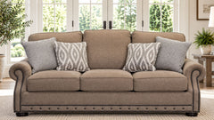 A three-seat upholstered loveseat in a warm taupe-beige fabric, styled with four throw pillows (three solid taupe and one gray-and-white geometric patterned pillow). The loveseat features dark wood nailhead trim along the base and rolled arms, set in a bright, modern living room with large windows and plants.