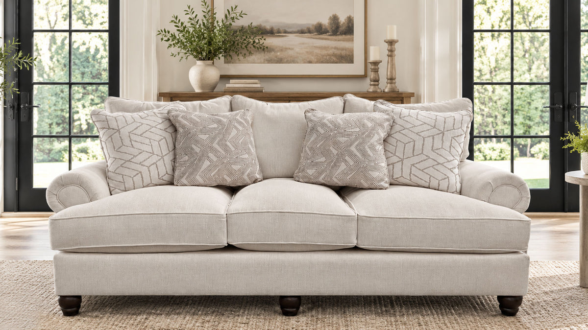 A light beige upholstered loveseat with rolled arms, dark wooden legs, and four decorative throw pillows, shown in a bright living room setting.