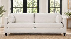 A light beige upholstered loveseat sofa with two back cushions, two seat cushions, and two matching throw pillows, shown in a bright living room setting with black-framed windows and a woven rug.