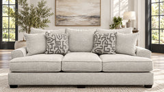 A light gray upholstered three-seat sofa in a bright living room, styled with three patterned throw pillows. The room features a neutral-toned rug, a side table with a lamp, a potted plant, and a large abstract painting on the wall.