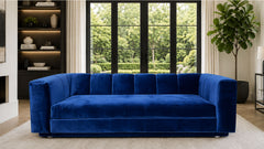 A modern blue velvet sofa with a tufted backrest, positioned in a contemporary living room with large windows showing a green garden view.